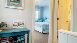 A hallway leading to a bedroom with a bed, a framed picture of a beach, and a sign on a table.