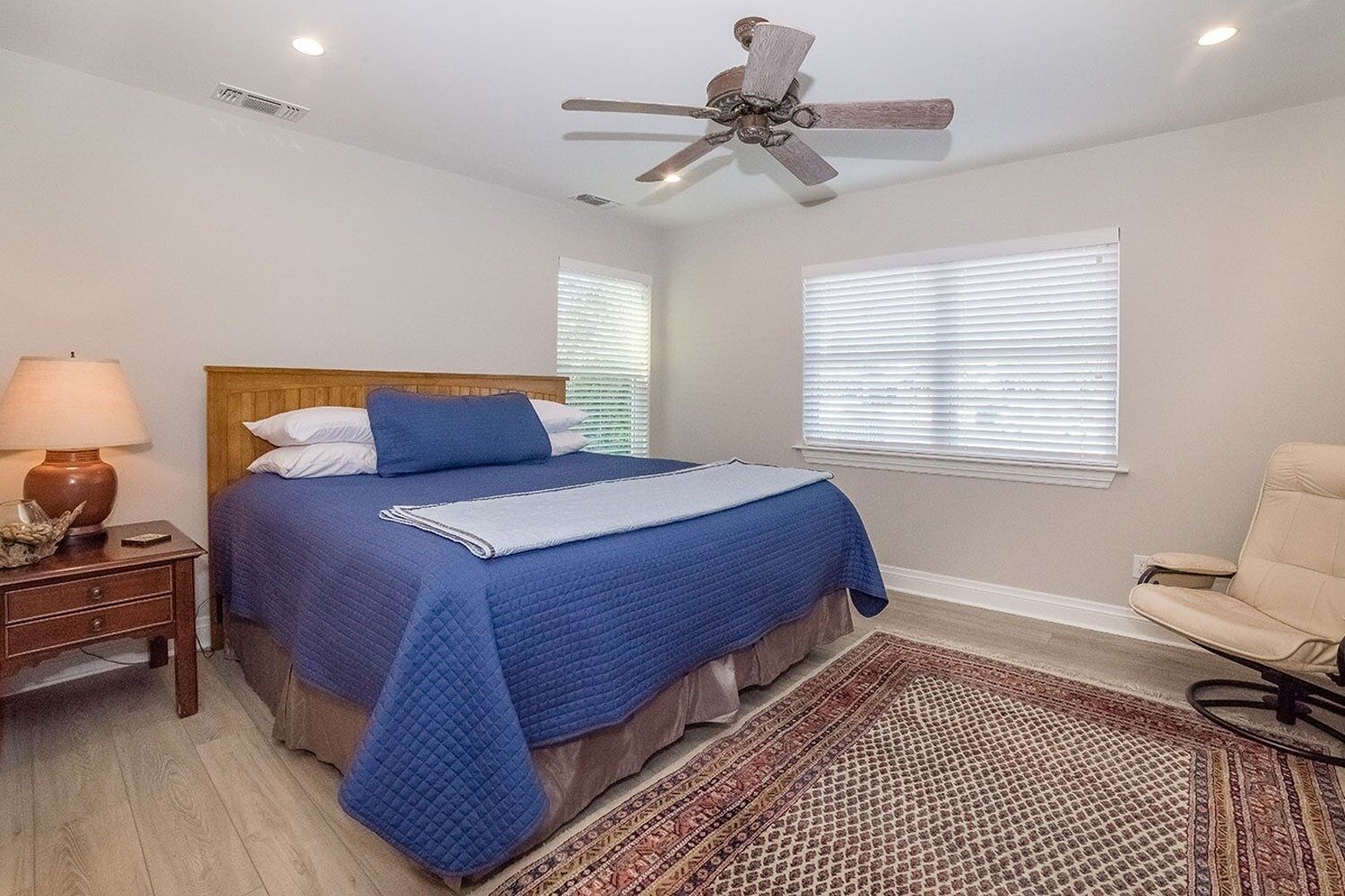 A bedroom with a bed, a ceiling fan, a nightstand, a lamp, a chair, and a window with blinds.