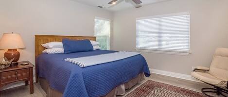 A bedroom with a bed, a ceiling fan, a nightstand, a lamp, a chair, and a window with blinds.