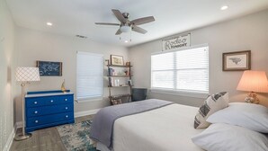 A bedroom with a bed, a blue dresser, a bookshelf, a ceiling fan, and two windows with blinds.
