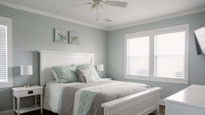 A bedroom with a white headboard, a bed with a quilt, two bedside tables with lamps, a nightstand, a window with blinds, and a ceiling fan. - Discover coastal charm at Holden Pattern, a spacious 4BR home in Holden Beach. (Holden Beach)
