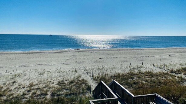 A beach with a wooden structure in the foreground, a clear sky, and the ocean in the background.