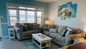 A living room with two grey sofas, a white coffee table, a wooden dining table, and a green patterned rug.