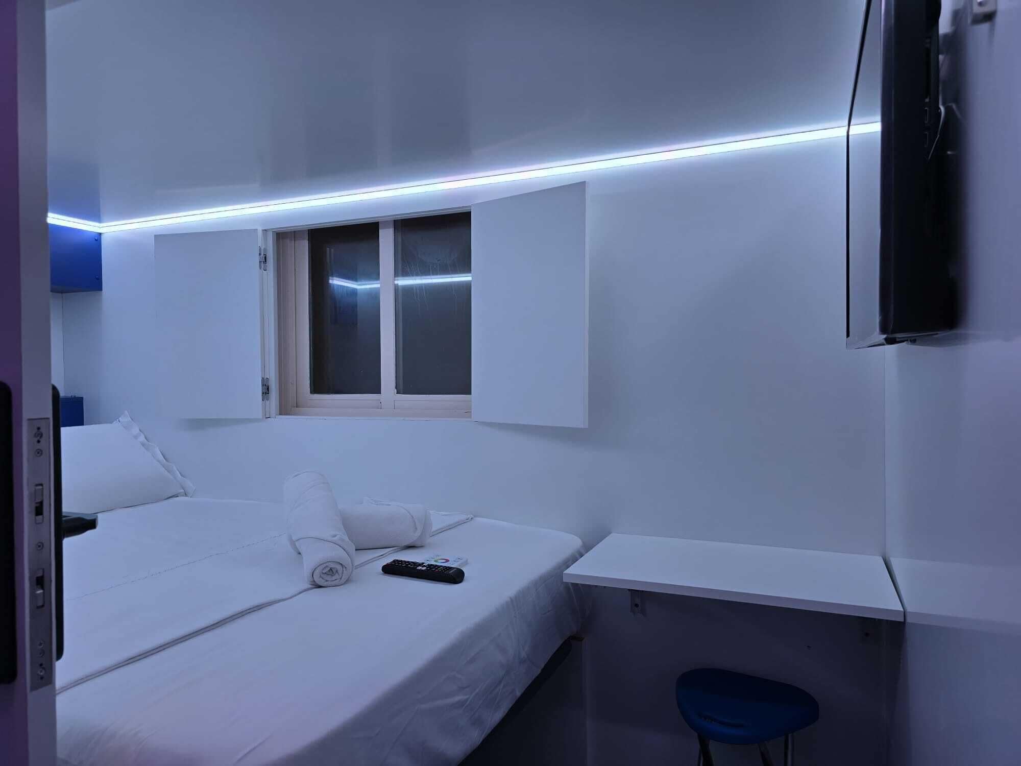 A hotel room with a bed, a desk, a chair, a TV mounted on the wall, and a window with blinds.