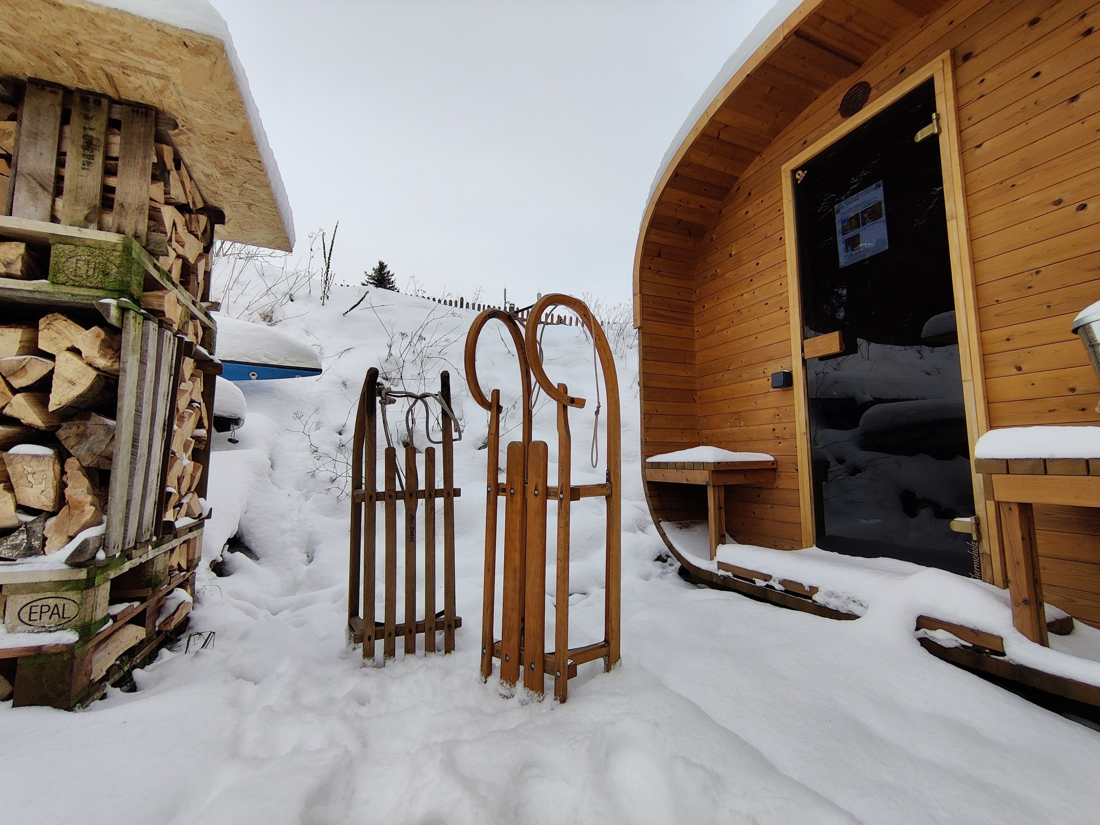 Een houten blokhut met een besneeuwde ingang, een houtstapel en een bank ervoor.