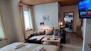 A hotel room with a bed, a sofa, a small table, a TV, and a kitchen area in the background.