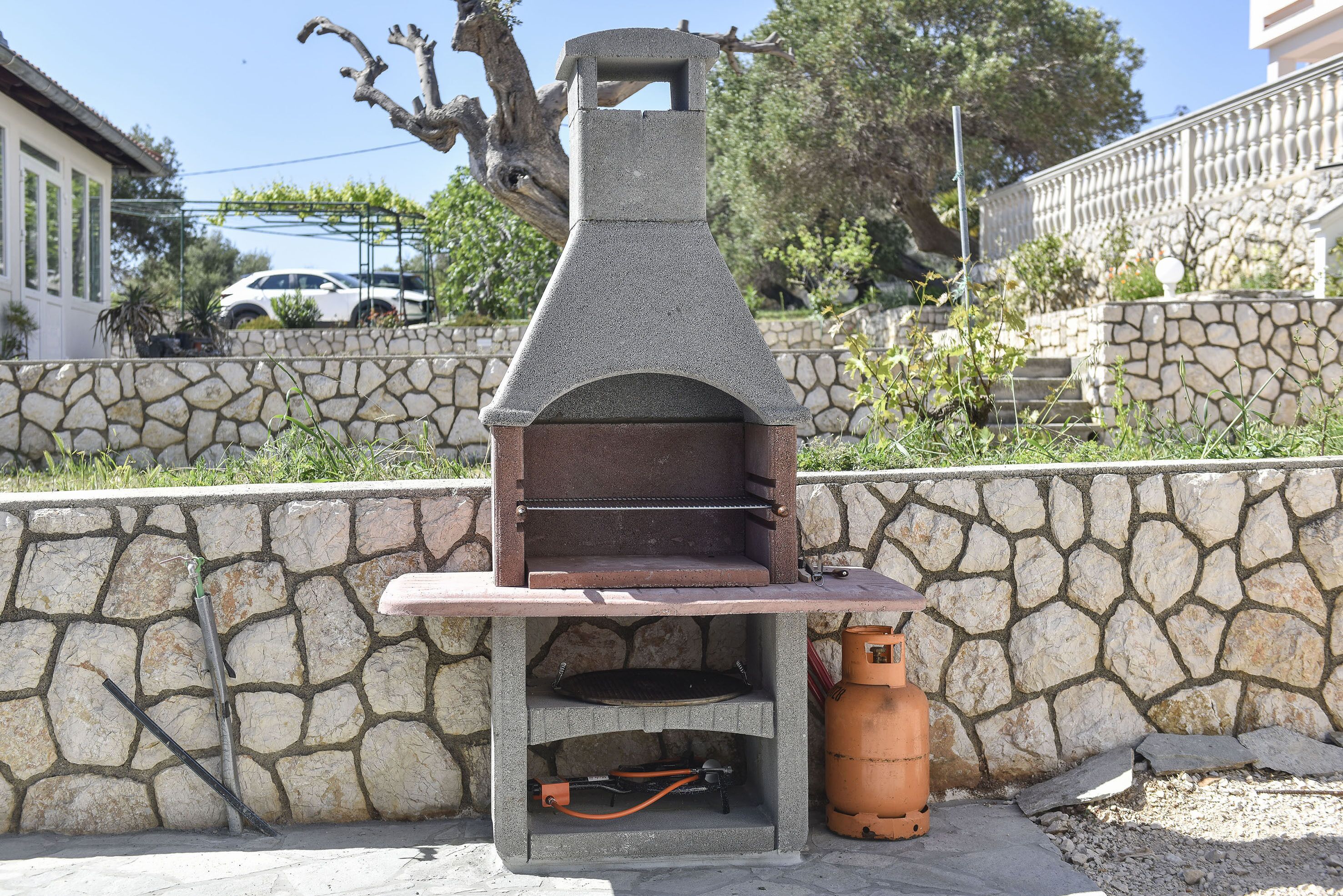 Un barbecue extérieur en pierre, doté d’une cheminée, d’une bouteille de gaz et d’une étagère en métal.