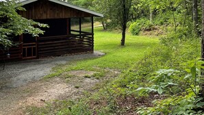 Una cabana rústica amb un terrat inclinat i una terrassa de fusta, envoltada per un bosc dens i una zona verda.