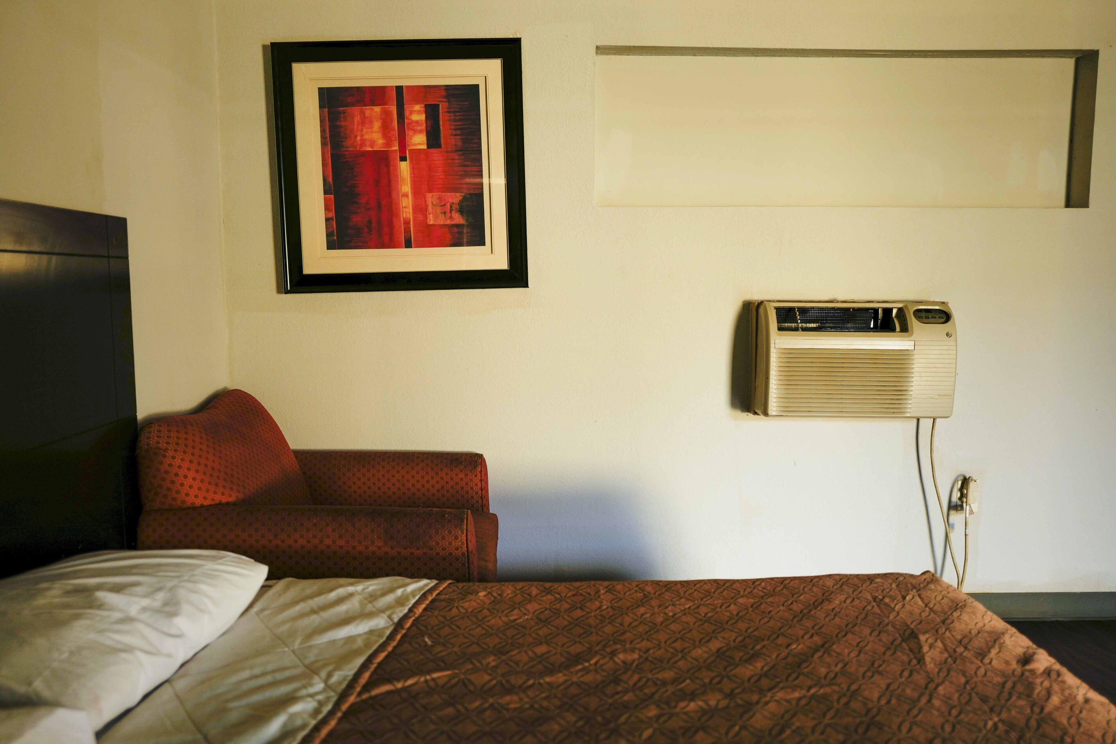 A hotel room with a bed, a red sofa, a framed artwork on the wall, and an air conditioning unit.
