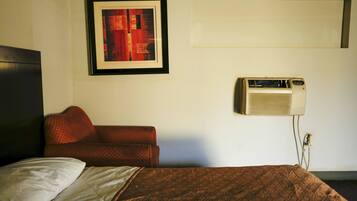 A hotel room with a bed, a red sofa, a framed artwork on the wall, and an air conditioning unit.