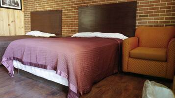 A hotel room with a wooden floor, a large bed with a headboard, a brown headboard, a patterned orange armchair, and a white trash bin.