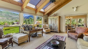 Un salon spacieux avec un plafond haut, de grandes fenêtres offrant une vue sur les montagnes, une cheminée chaleureuse et un coin repas avec vue.
