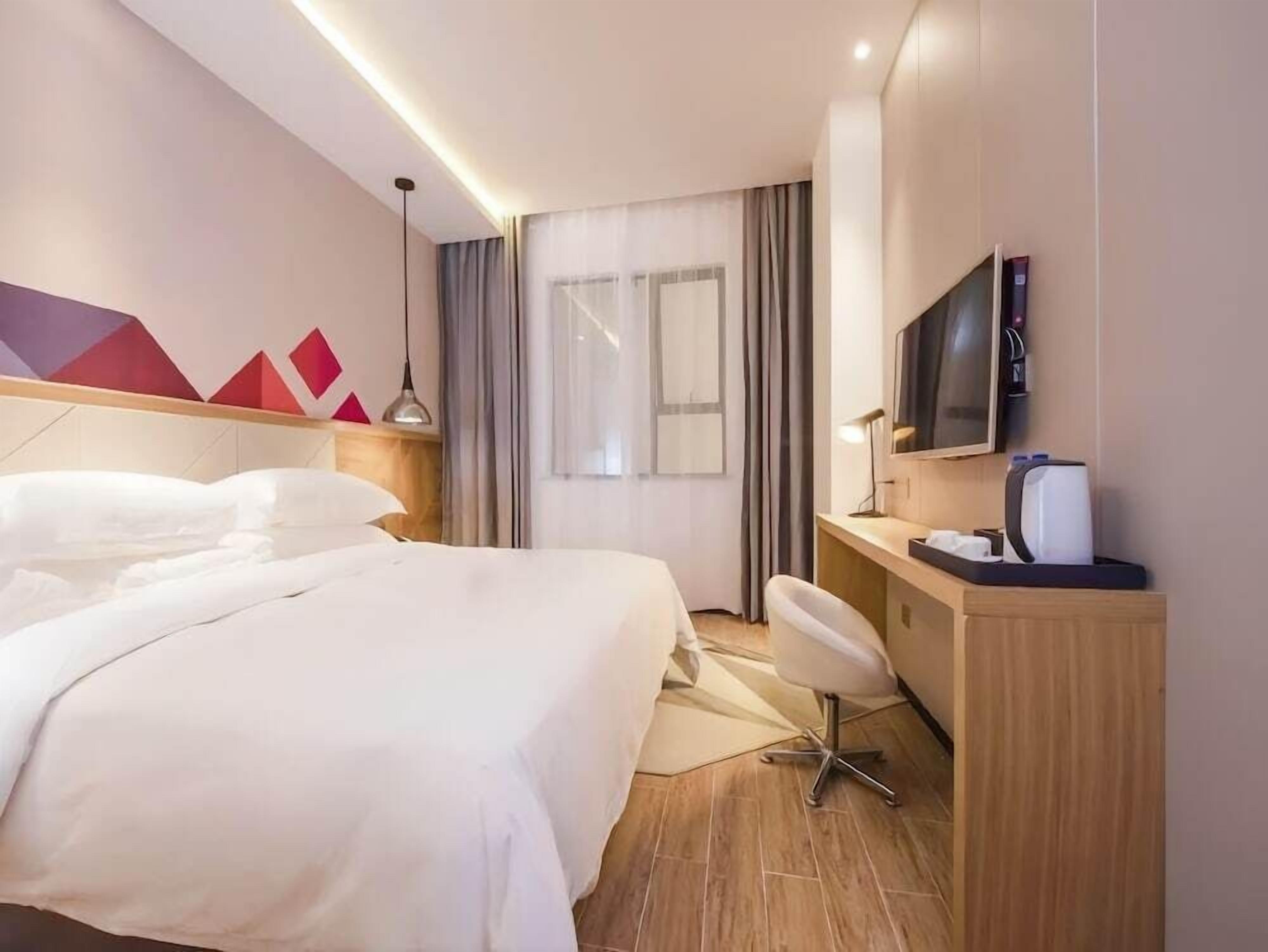 Ein Hotelzimmer mit einem großen Bett, einem Schreibtisch mit Stuhl, einem Fernseher und einem Fenster mit Vorhängen.