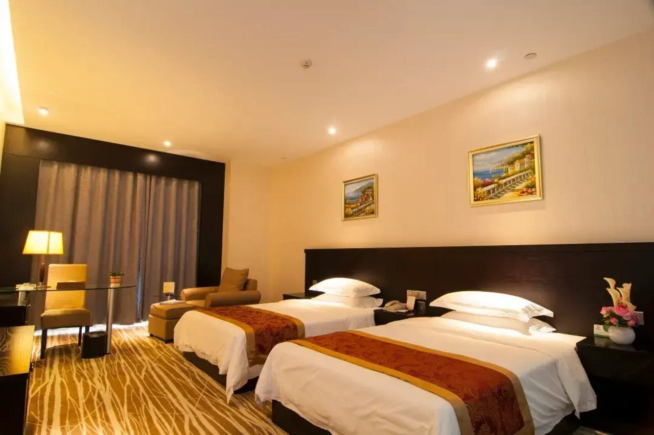 A hotel room with two beds, a desk, a chair, a lamp, and a painting on the wall.