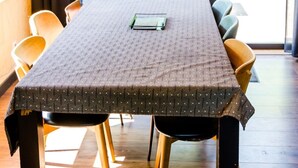 Una mesa de comedor con mantel estampado, rodeada de sillas, en una habitación con ventanas amplias y una lámpara colgante.