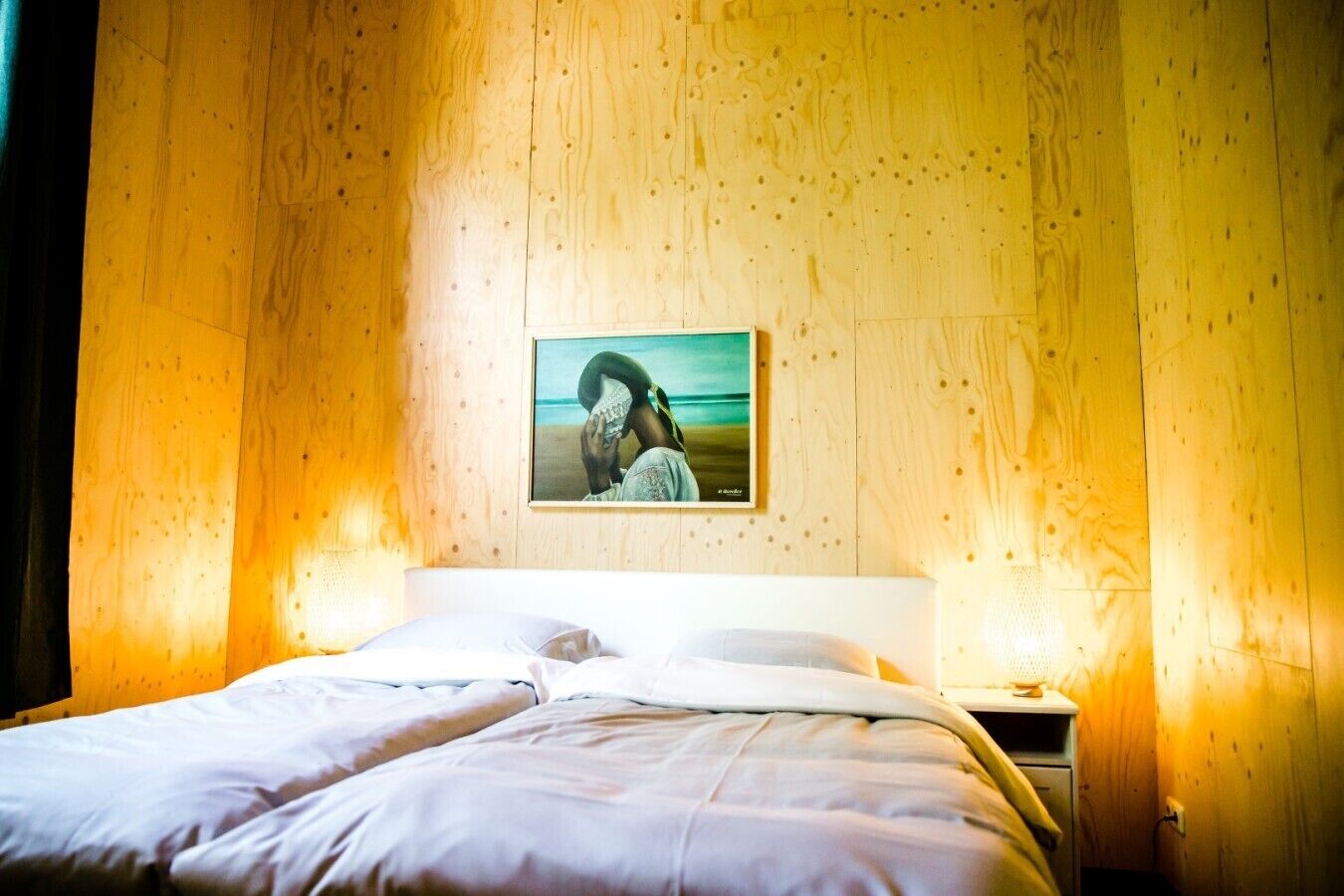 Uma cama com lençóis brancos, mesa de cabeceira com abajur e quadro na parede.
