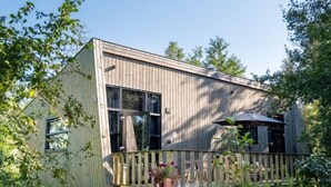 A wooden cabin with a deck, surrounded by tall grass and trees.