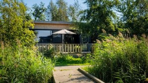 A wooden boardwalk leading to a modern building with a glass facade, surrounded by lush greenery and a clear blue sky. - Nature lodge Reade Wikel, Akkrum - children`s sleeping hut (Akkrum)