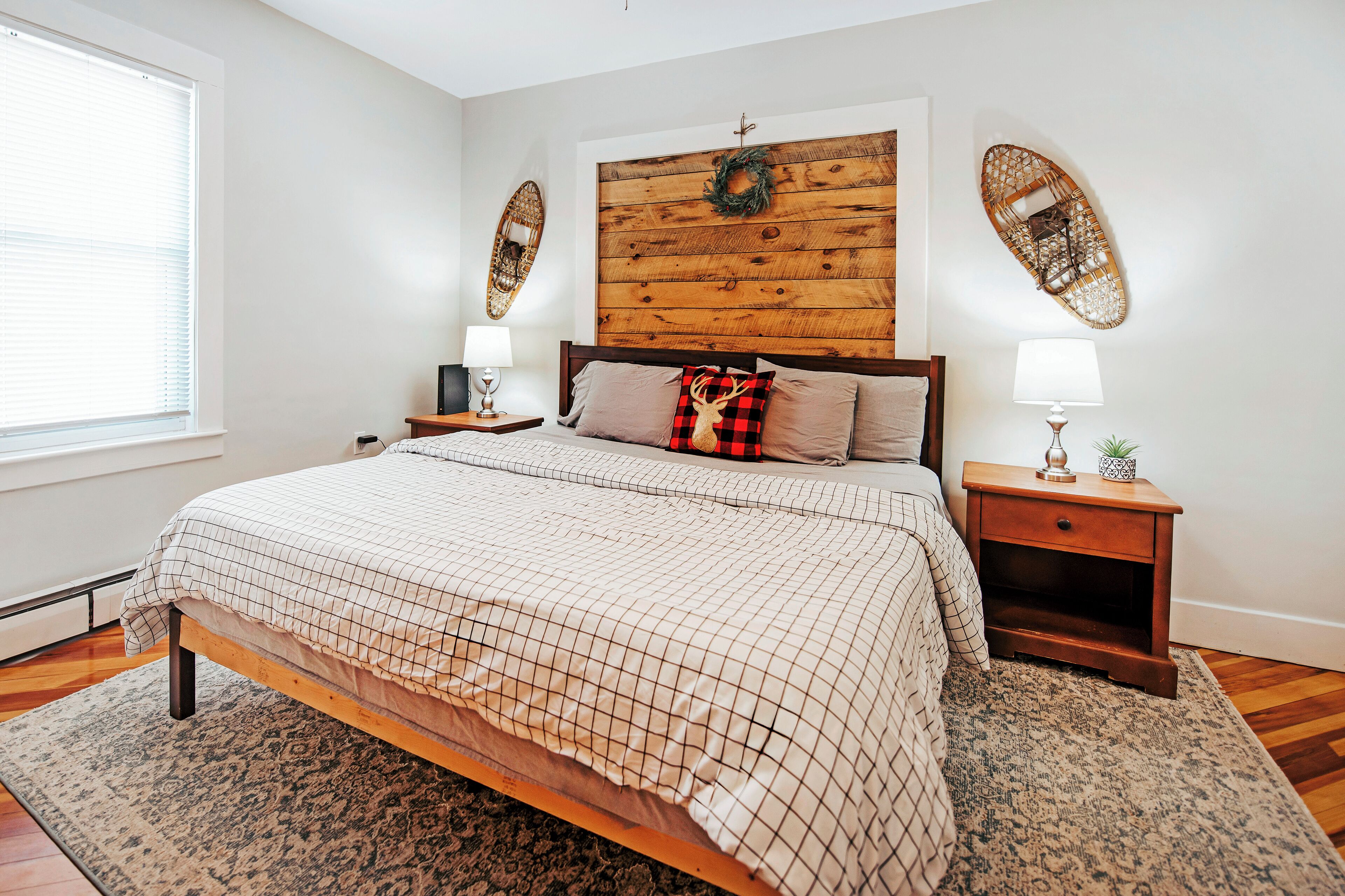 Un dormitorio con cabecera de madera, cama con colcha a cuadros, dos mesitas de noche con lámparas y un ventanal con persianas.