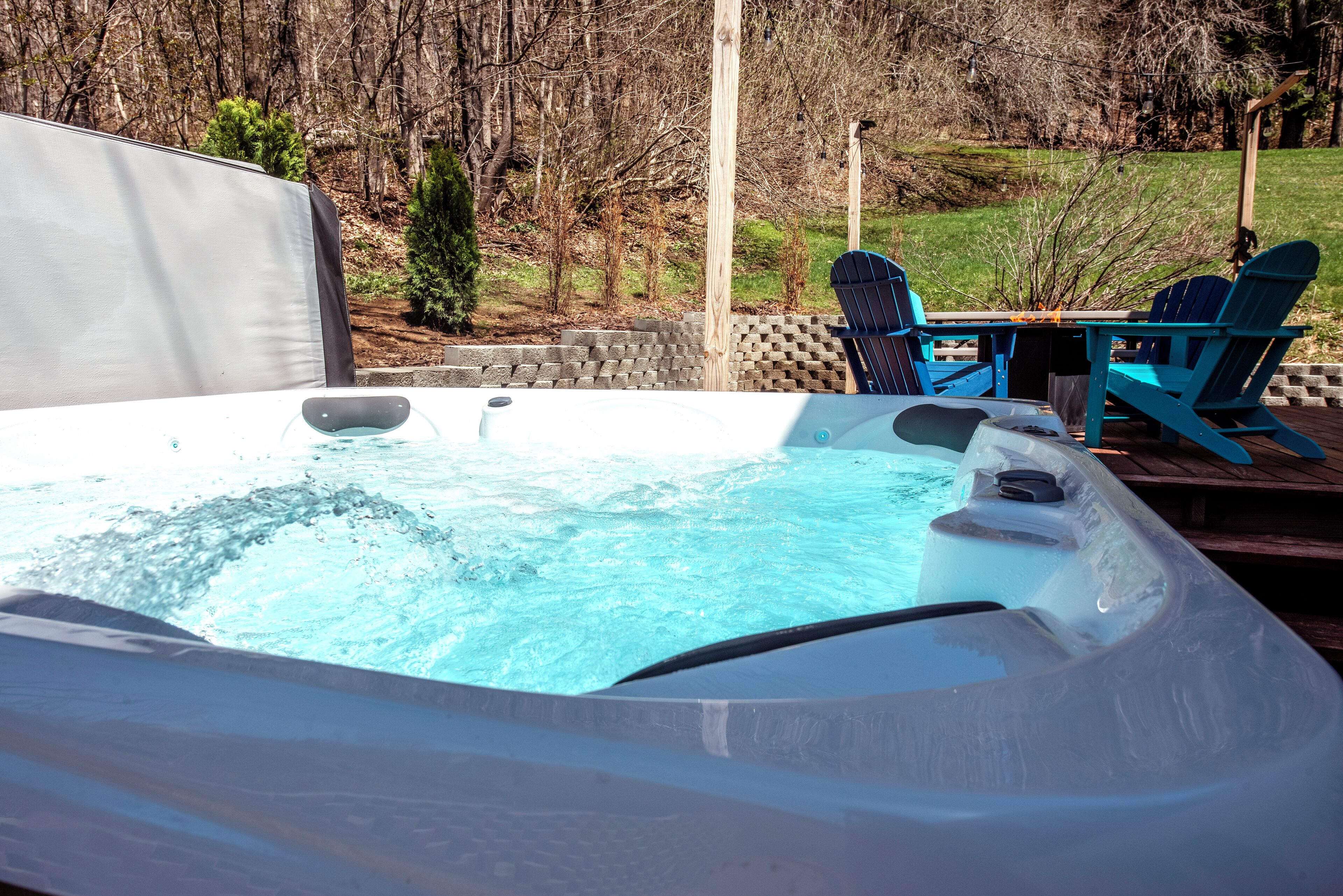 A hot tub with a wooden deck and chairs, surrounded by a stone wall and trees.