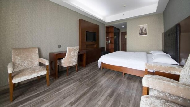 A hotel room with a bed, two armchairs, a desk, a television, and a bookshelf. - Sicily Hotel (Changsha)