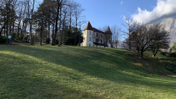A castle with a steeply pitched roof and a tower, surrounded by a grassy area with trees.
