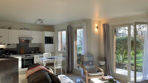 A modern living room with a sofa, a coffee table, and a dining area with chairs. There is a kitchen area with cabinets and appliances. The room has large windows with curtains, providing a view of the outdoors.