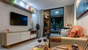 A modern living room with a flat-screen TV, wooden furniture, a dining table with a bottle of wine, and a view of the outdoors. - Apto térreo com TV 4k no Wai Wai Cumbuco (Caucaia)