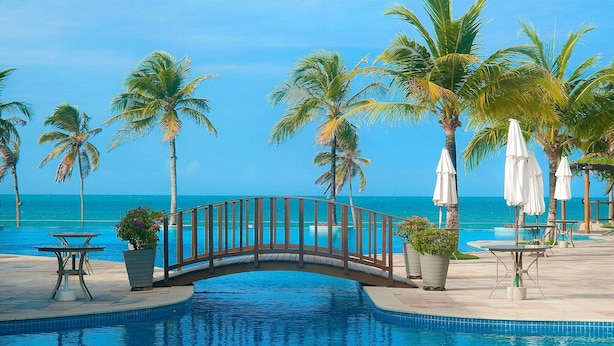 A pool area with a bridge, palm trees, and a view of the ocean. - Apto térreo com TV 4k no Wai Wai Cumbuco (Caucaia)