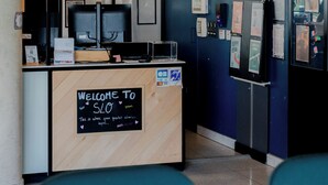 Een receptiebalie met een bord waarop 'Welcome to SLO' staat, een computer, ingelijste foto’s aan de muur en een blauwe muur met diverse mededelingen.