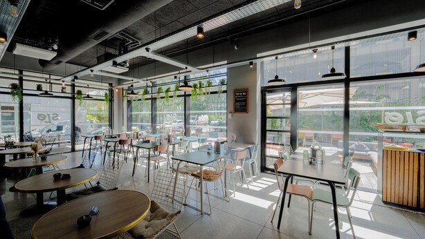 A modern cafe interior with tables and chairs, large windows, and hanging plants. - Slo Bordeaux Hostel (Bordeaux)