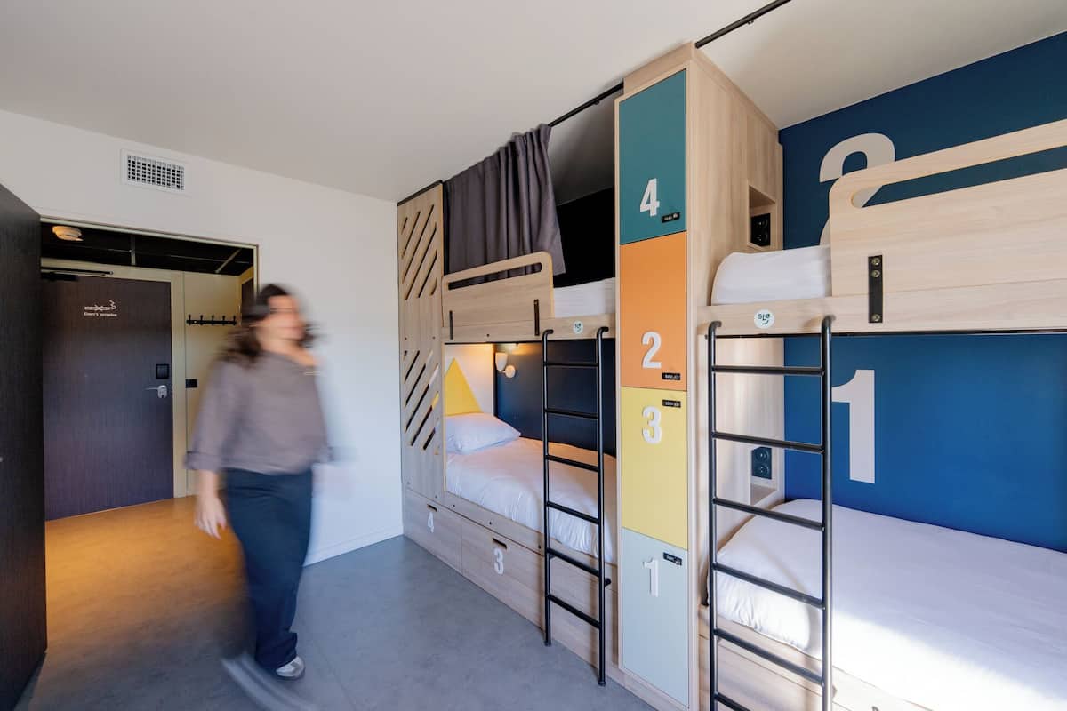 Une chambre avec des lits superposés, des numéros indiquant l’attribution des chambres, une porte sur laquelle est affiché un panneau « Il est interdit de fumer », et une personne qui passe.