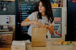 Une personne se tient derrière un comptoir, et on aperçoit en arrière-plan un panneau affichant le menu avec les plats proposés et leurs prix.