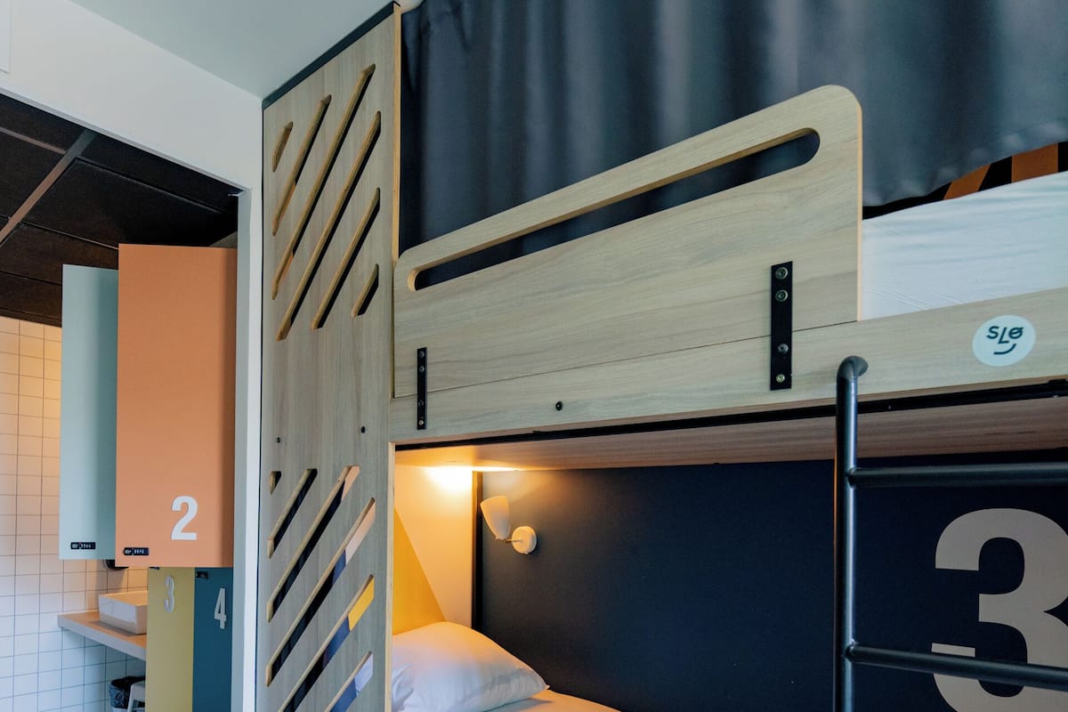 Un lit superposé avec une tête de lit en bois, un bureau et une chaise, dans une pièce dont le sol est carrelé.