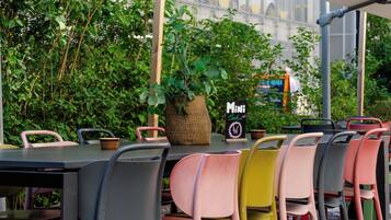 An outdoor seating area with tables and chairs, a signboard, and greenery in the background.