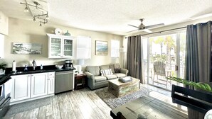 A modern living room with a sofa, coffee table, and a dining area with a table and chairs. There is a large window with curtains, a ceiling fan, and a kitchen area with cabinets and a sink.