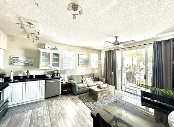 A modern living room with a sofa, coffee table, and a dining area with a table and chairs. There is a large window with curtains, a ceiling fan, and a kitchen area with cabinets and a sink.