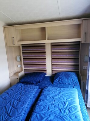 A bedroom with two beds, a headboard with striped cushions, a wooden cabinet, and a small shelf.