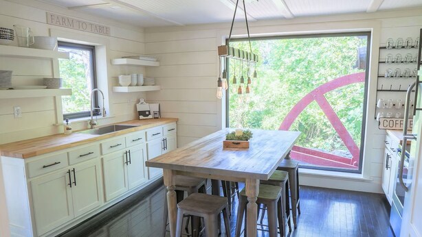 Una cocina con armarios blancos, encimera de madera, zona de asientos con taburetes de bar y un amplio ventanal con vista a la vegetación.