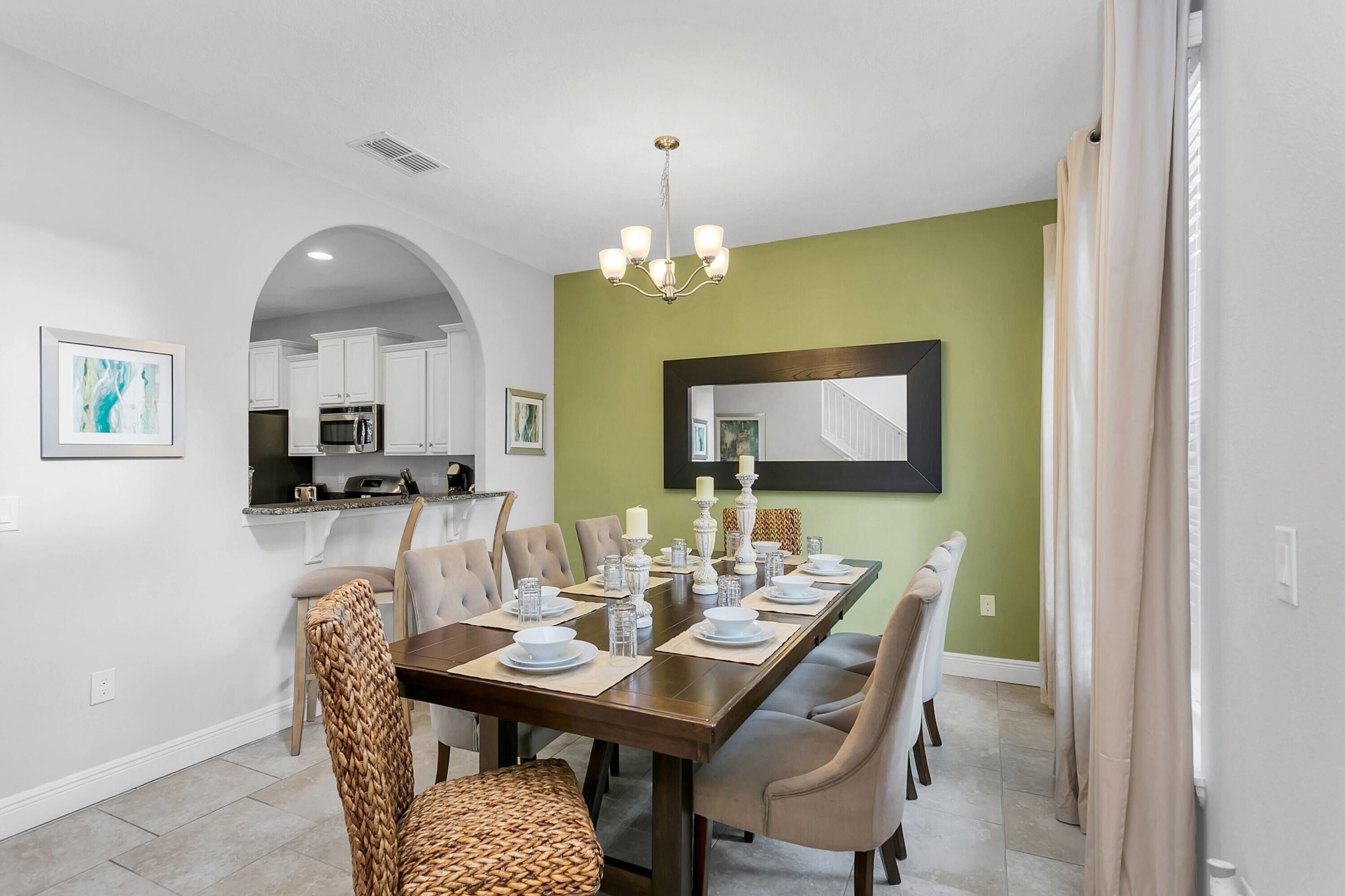Uma sala de jantar com uma mesa comprida posta para seis pessoas, um candelabro e uma cozinha ao fundo.