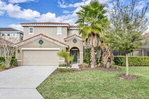 A two-story house with a beige facade, a garage, and a well-maintained lawn with palm trees.
