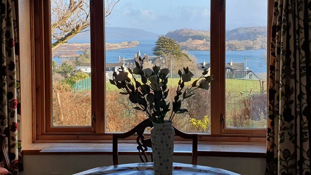 A room with a large window offering a view of a coastal landscape, a wooden chair, and a vase with flowers on a table. - An Cala - Spacious house with stunning sea-views (Tobermory)