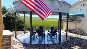 Eine überdachte Terrasse mit Blick auf ein grünes Feld, mit einem Pavillon, Gartenstühlen und einer amerikanischen Flagge.