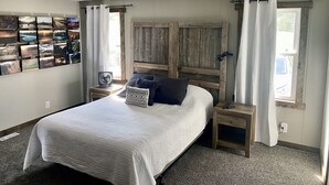 Ein Schlafzimmer mit einem Bett, einem Holz-Kopfteil, einem Nachttisch mit Lampe, einem Wandventilator und einem Fenster mit Vorhängen.