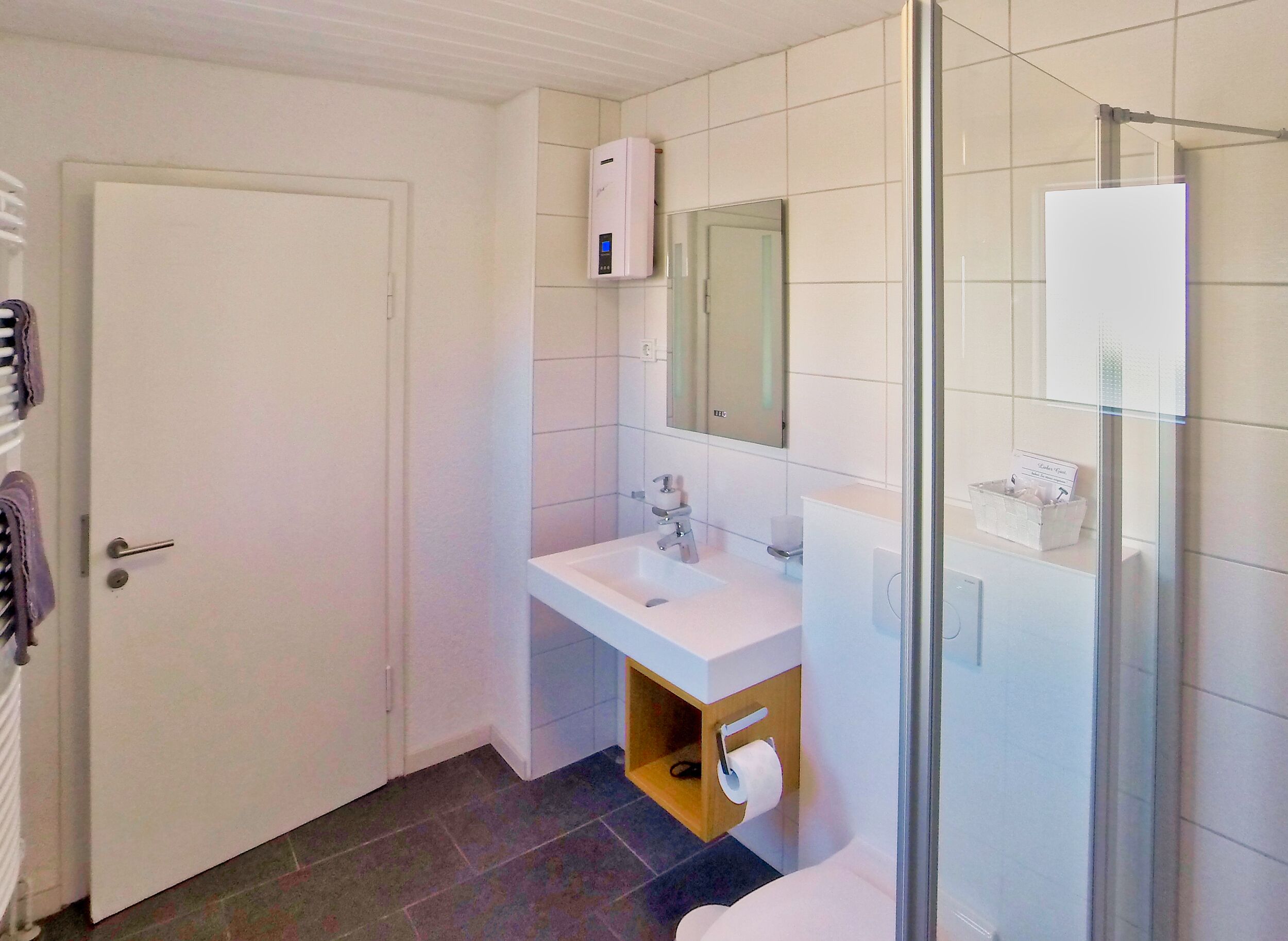 A bathroom with a white sink, mirror, and toilet. There is a shower area with a glass door. A towel rack is visible on the left side.