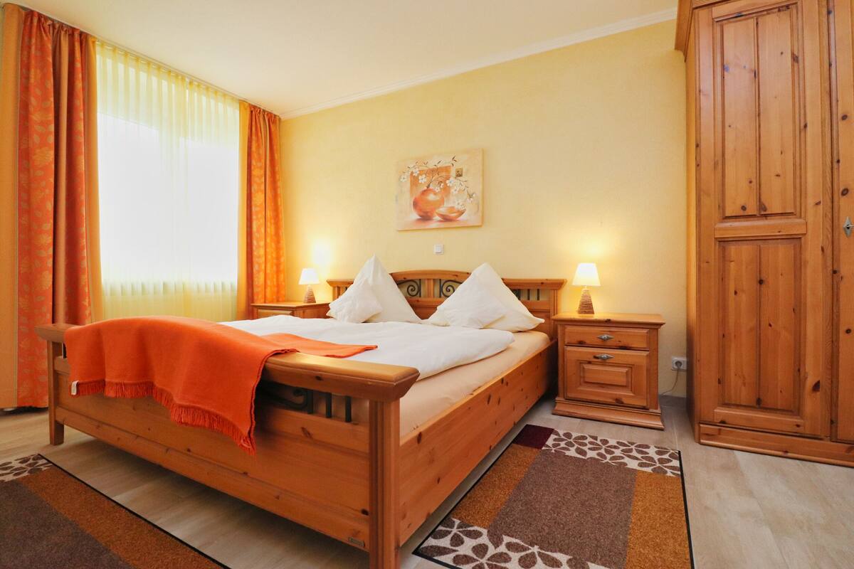Een houten bed met wit beddengoed, twee nachtkastjes met lampen, een houten garderobe en een schilderij aan de muur.