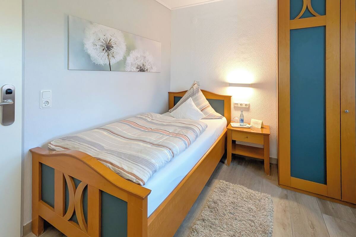 Een enkel bed met houten frame en een gestreept laken, een nachtkastje met een lamp, een wandlamp en een deur met een blauw paneel.