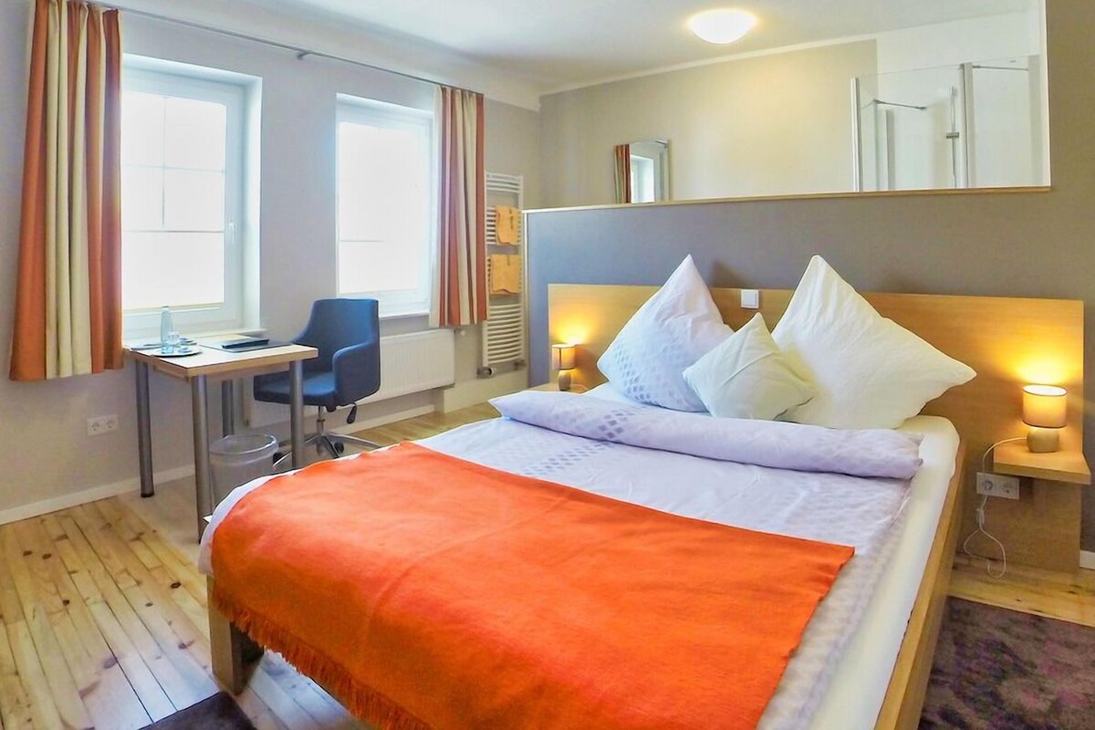Een hotelkamer met een bed, een bureau en een stoel. Er zijn ramen met gordijnen, een spiegel en een lamp.