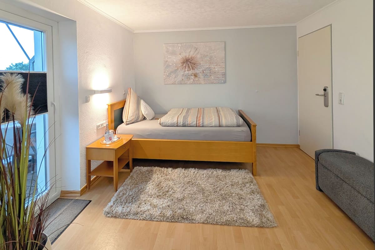 Een kleine, knusse slaapkamer met een eenpersoonsbed, een nachtkastje met een lamp, een grijze bank, een pluizig vloerkleed en een groot raam met uitzicht.