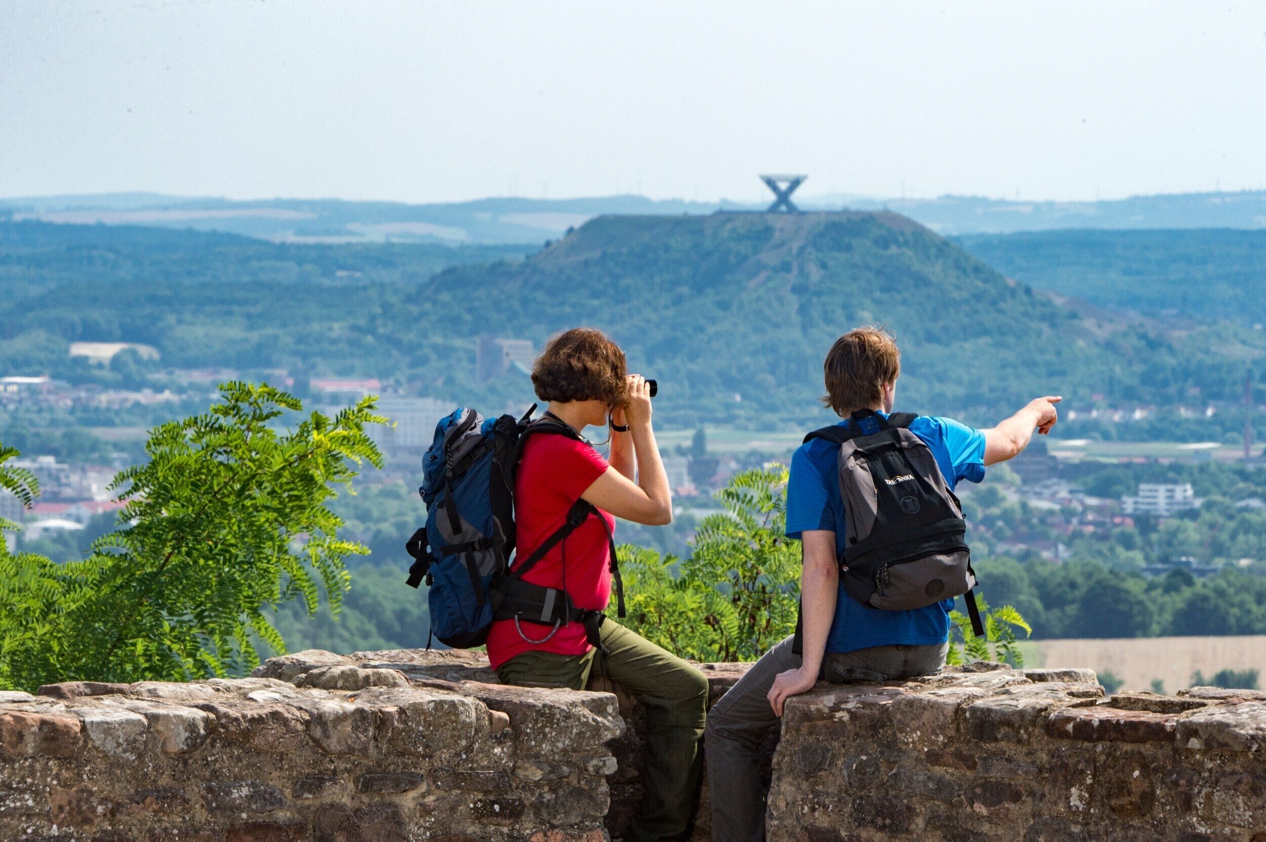 Dos personas sentadas en un muro de piedra con vistas a una ciudad y una colina con una estructura distintiva.